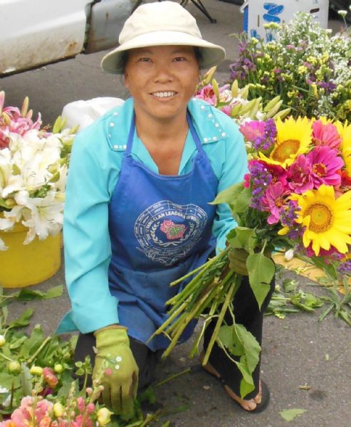 Hmong gardener kneeling and looking into the camera at the Northside Farmer’s Market. She is wearing a white hat, light blue shirt, royal blue apron, green gloves, black pants and black sandals. On the apron is the logo for "The 12 Yang Clan Leaders of Wisconsin, Inc." In her left hand is a bouquet of flowers, and flowers in buckets surround her.