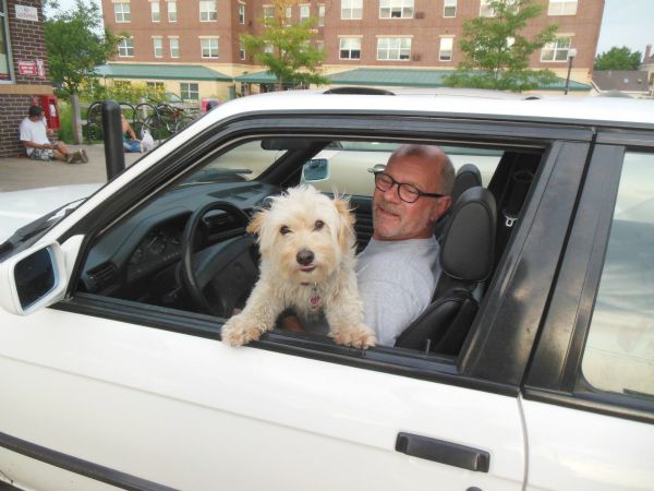 Bob Monk sits behind the wheel of his car, holding his fluffy dog in his lap, in the Kwik Trip parking lot. The dog has its paws on the edge of the car door as it peers out at the photographer. In the background is a large building and trees, on the left are bicycles and people sitting on the ground.