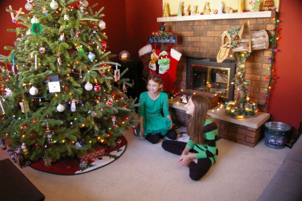 Two teen girls gaze at a Christmas tree with gifts underneath as they sit on the floor in the Temp home. One girl is wearing a turquoise dress and black tights, the other girl wears a green and black striped sweater with black leggings. Behind them is a fireplace decorated with lights. On the left are four stockings, on the right is a mailbox for holiday cards and on the mantle is a Nativity scene.