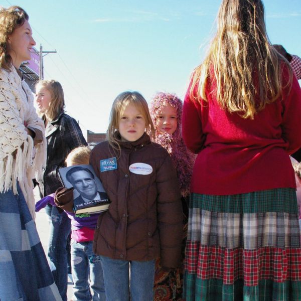 A girl wearing a coat with patches for Tommy Thompson for Senator and Feehan for State Senator stands holding a Mitt Romney book. Her sister stands behind her. They are outside of the Paul Ryan Election Rally at the La Crosse County Republican Headquarters. Their mother has her back to the camera. They are surrounded by other children and women.