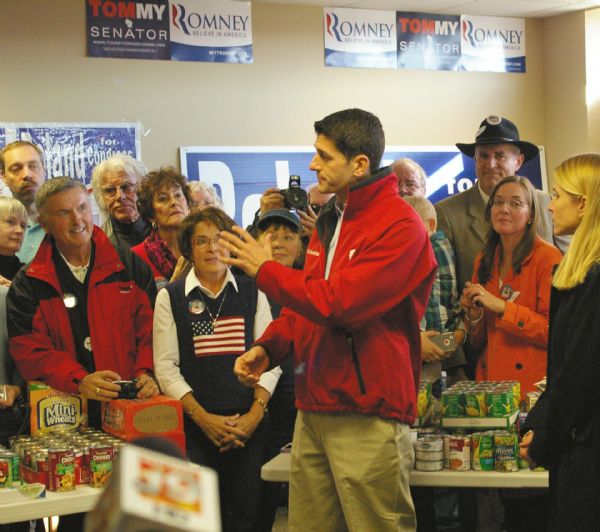 Congressman Paul Ryan speaks to a group as the Republican Vice Presidential candidate at the La Crosse County Republican Headquarters. Most people are wearing coats and campaign pins. One women is wearing a sweater decorated with the American flag. Non perishable food items cover two tables. Campaign posters are hanging on the walls.