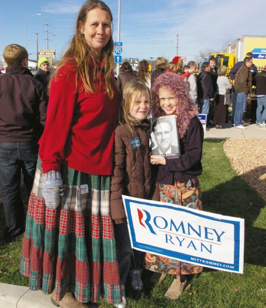 A mother and two daughters pose outside of a Paul Ryan Rally Event at the La Crosse County Republican Headquarters. One girl holds a Mitt Romney book. The mother is wearing a long patchwork skirt and sweater with mittens, and the girls are wearing coats. The girl on the right is wearing a skirt and a fluffy scarf. A Romney-Ryan sign is in the foreground. People are lined up in the background.