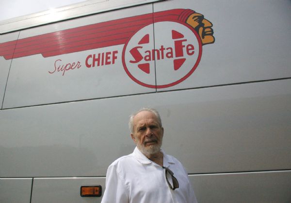 Merle Haggard posing in front of his Super Chief Santa Fe motor coach at the Southside Oktoberfest Grounds. He has his sunglasses hooked in the collar of his white shirt.