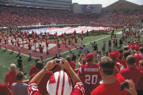 A giant American flag is spread across the field by marchers as the marching band plays, before Gary Andersen’s first football game as UW-Madison head coach. The Badgers played the UMass Amherst Minutemen at Camp Randall Memorial Stadium. In the foreground are Badger fans, many with their cameras over their heads taking snapshots.