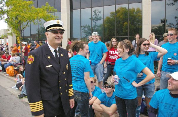 La Crosse Fire Chief Greg Cleveland, standing along 2nd Street, at the Oktoberfest Parade. He is wearing his dress uniform and sunglasses. On the sidewalk, various groups wear identical t-shirts. The blue shirts display a cartoon man with a bratwurst.