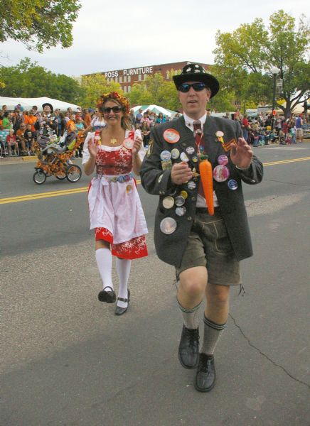 Oktoberfest frolickers on parade during the Mapleleaf Parade on 2nd Street. Carie Ann (Schlichenmeyer) Finch and man in the foreground are wearing ethnic costumes with many buttons attached. The man has pins on his hat and a carrot on a ribbon around his neck. A decorated stroller is behind them on the left. Spectators line the curb. In the background are tents, trees and the Ross Furniture Company building.