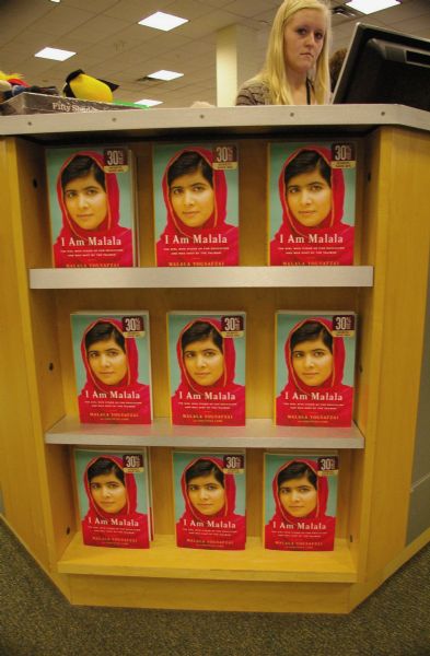 The book “I Am Malala” on display beneath the sales counter at Barnes & Noble Booksellers. A sales clerk with blonde hair is looking down from her position at the cash register.