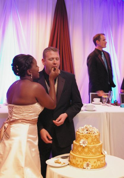 Unidentified bride and groom with wedding cake. The bride is feeding a slice of wedding cake to the groom. The cake is frosted in gold with white flowers and orange piping. She is wearing a strapless gown with laces in the back and he is wearing a black tuxedo. In the background is the wedding table with tableware, and another member of the wedding party is standing and wearing a tuxedo in front of red and lavender drapes.