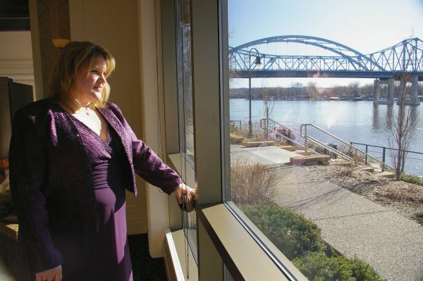 The owner of La Crosse Radio Group, Jacklyn Daniels, gazes out of the window at the riverside. The event was the DMI Trend Showcase in the  Cargill Room. She is wearing a purple dress with a matching jacket. The view out of the window includes a sidewalk, steps down to the Mississippi River, and a bridge.