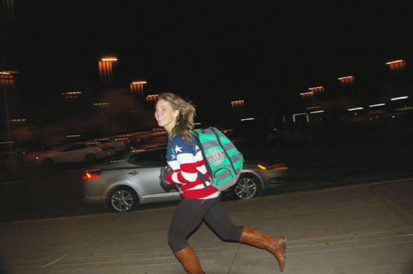 A young woman wearing an American flag design sweater, with a green and grey striped knapsack, skateboarding (skateboard unseen) on the sidewalk outside of the Soledad O’Brien event at UW-La Crosse. It is after dark. Automobiles and lights are in the background.