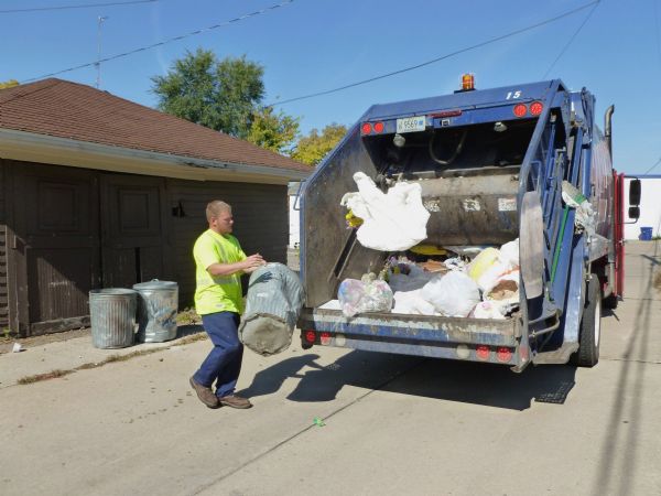 The driver of a Harter’s Refuse truck throws a bag of trash into his truck. He is holding one trash can, and two others sit near a garage. He is wearing a safety green t-shirt with reflective stripes, blue pants and work boots.
