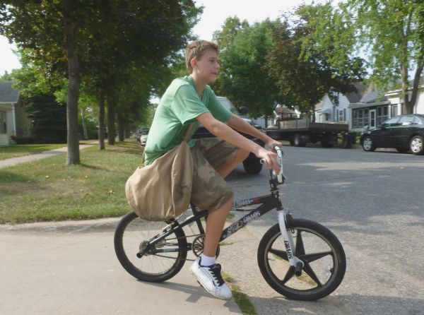 Paper boy riding a bike along Charles Street on Labor Day. He is wearing a green shirt, camouflage shorts and white tennis shoes. In the background is a neighborhood with sidewalks, trees and parked automobiles.