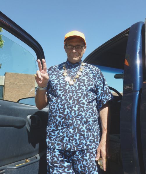 A colorfully dressed man flashing a peace sign while in town attending a designers’ conference. An ornate necklace is hanging around his neck and he is wearing a baseball cap. He is parked in the La Crosse Public Library parking lot.