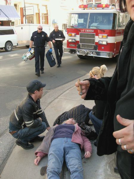 A woman is lying unconscious on the sidewalk, with two people at her side, on the corner of 4th & Pearl Streets. The Heavy Rescue fire truck has arrived with emergency personnel. In the foreground on the right, a man holding a cigar is partially visible.