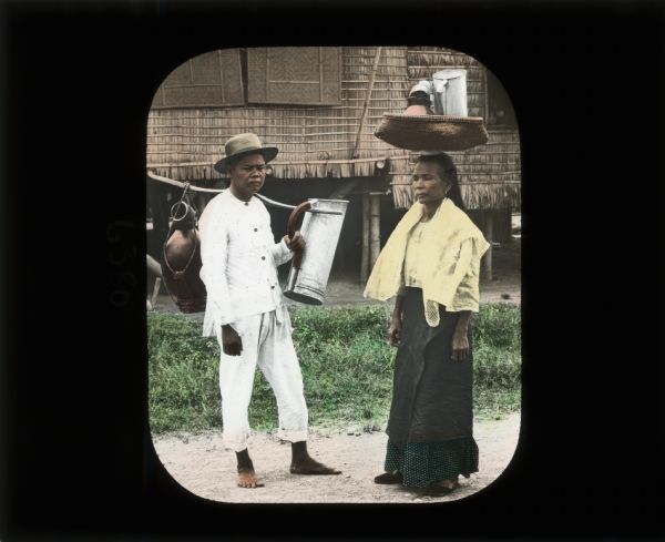 A man and woman gathering water in calabashes, pitchers and clay pots. The women is carrying the water on a basket on her head, while the man carries them on a pole over his left shoulder. They are standing in front of a common type of housing made from woven fibers. The man is wearing white clothing and a hat, and is barefoot. The woman is wearing a yellow blouse, green skirt, and slippers.
