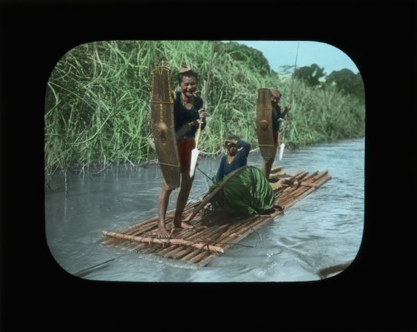 Three people on a raft, all wearing blue shirts. Two of the people are standing and holding shields and spears, while the third person is sitting in the middle near a large leaf shading their eyes from the sun. The person standing at the front of the raft has a large weapon sheathed at their waist, and has what may be a cigar in their mouth. In her journal from South Africa Carrie describes the "shocking blue" of the Zambezi River and the wide variety of boats, including "primitive rafts made of logs gathered together."