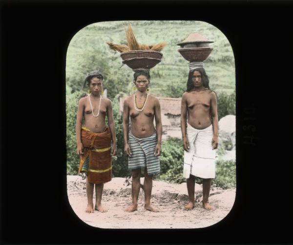 Three women standing in a row. Two of the women are carrying food in baskets and pottery on their head. They are unclothed above the waist and wearing cloth tied around their waists as skirts, which was common practice in Ceylon (now Sri Lanka) at the time. They are all barefoot. The first two appear to have pierced ears and are wearing large earrings as well as necklaces. In the far background are buildings and a steep hill. In her journal from Ceylon Carrie records: "Both men and women wear a sort of skirt made of a straight piece of cloth wound around the body."
