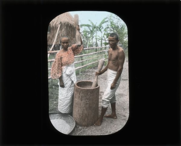 A woman and man grinding a variety of grain in a large stone mortar. The woman is wearing a blouse and skirt, the man is wearing white pants and is barefoot. Behind them is a fence and a house on stilts.