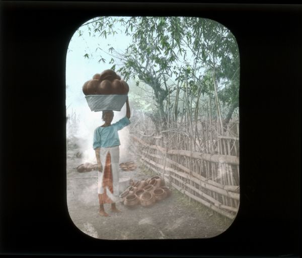 Full-length portrait of a woman standing and holding a basket full of pottery on her head. There is more pottery in a pile on the ground near a bamboo fence. The woman, who is barefoot, is wearing a blue blouse and an orange and white skirt.