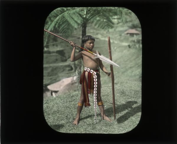 Portrait of a boy standing barefoot outdoors in the grass holding a spear in his right hand and what may be a shield in his left hand. He also has an implement of some type hitched into the belt around his waist. He is wearing red and gold clothing, and his spear is also colored in red and gold. Carrie, in her journal from her trip to Java, describes his belt as "disk-like beads which are strung to form a long, single strand."