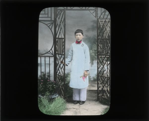 Hand-colored portrait of a boy standing in the arch of a decorative iron gate holding the door open with his right hand and holding a hand fan and handkerchief in his left hand. In the background is a painted backdrop of trees near a body of water. The boy appears to be wearing a uniform similar to that described by Carrie in her journal from Japan and South Korea which is worn by the boys who serve at temples. She describes the uniforms as "trousers, and worn above those a smock which has a collar fastened by a pin."