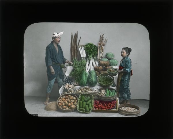 Hand-colored lantern slide of a man and a girl or young woman standing next to a market stand which has melons, roots, and other foods. The man is wearing a plain kimono with a white cloth wrapped around his head. The girl is wearing a patterned kimono.