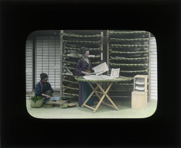 Hand-colored lantern slide of two women working. The woman on the left is kneeling on the ground and chopping tea on a wooden board. The woman standing in the center is working with paper on a table. The racks behind her are used for drying. Both women are wearing kimonos.