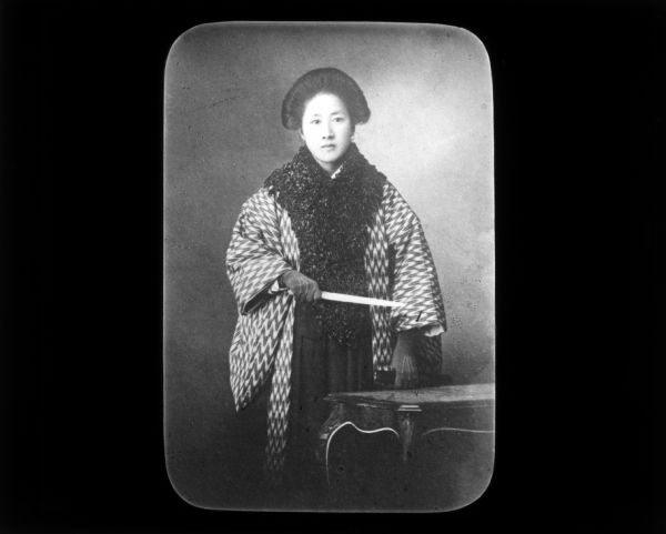 Three-quarter length portrait of Chinese revolutionary and feminist Qiu Jin in "European dress." She is standing near a table and holds an object in her right hand. Carrie Chapman Catt in her journals records that it is common for wealthy women to have their portrait taken in European clothing such as tailleurs (a women's suit which consists of matching jacket and skirt).