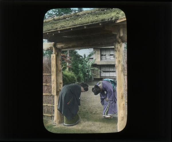 Hand-colored lantern slide of two women bowing to each other on opposite sides of an entrance gate to a yard. In her diary from Japan and South Korea, Carrie Chapman Catt describes the bowing as "a custom used for greeting, and for bidding farewell."
