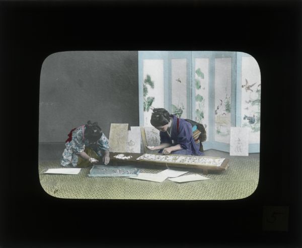 Hand-colored lantern slide of two girls in kimonos kneeling on a floor, bent over a low table working with paper. Behind them is a decorated folded room screen.