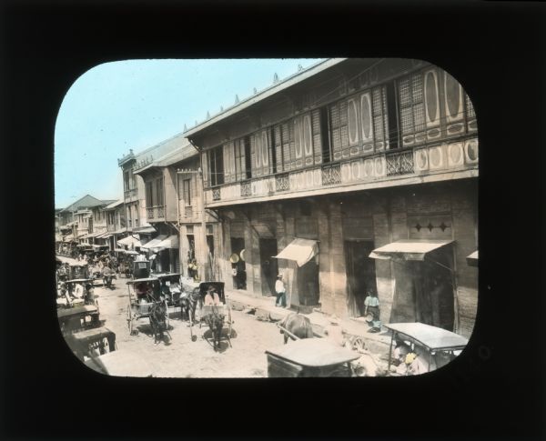 Hand-colored lantern slide of an elevated view of a street scene which includes an assortment of carriages. In her journal of Ceylon, Carrie Chapman Catt records that carriages are "most often pulled by a bullock, a sort of small cattle with a hump on its neck, and also by mules."