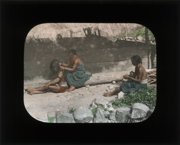 Four people sitting on the ground outside of a shelter with a thatched roof. The woman on the left is styling a girl's hair. The woman on the right is holding a baby. Carrie Chapman Catt in her journals often describes that many people in the countries she visits are unclothed above the waist.