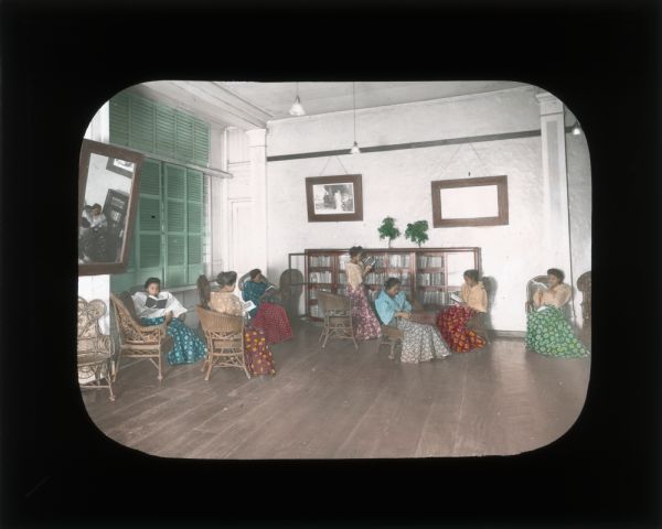 A group of women reading in a large room. One woman stands in front of a glass-fronted bookcase against the back wall. The woman wear brightly colored and patterned dresses and blouses with puffy sleeves. Carrie Chapman Catt's travels involved speaking to women about issues such as voting rights and wages. Her aim was to help establish political organizations for suffrage. In her journals she often describes the conditions of women in the area, including their literacy, social status, working conditions, and ownership rights.