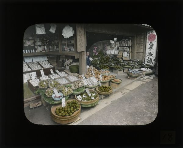 A market stall with an array of fruits, vegetables, fish, and flowers. The shop keeper is sitting in the center just behind a wooden post. In her journal from Japan and Korea, Carrie Chapman Catt describes the markets as "a marvelous opportunity to try any number of strange new things".