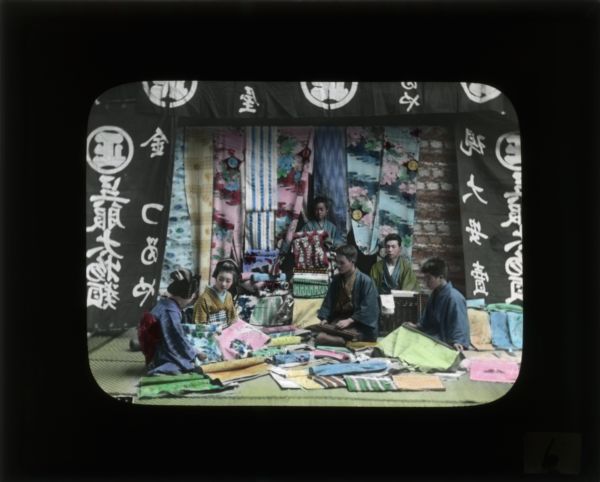 A group of young women in kimonos examining fabric while young men look on. In her journal from Japan and South Korea, Carrie records that fabric is one of the many things to be found at the markets.