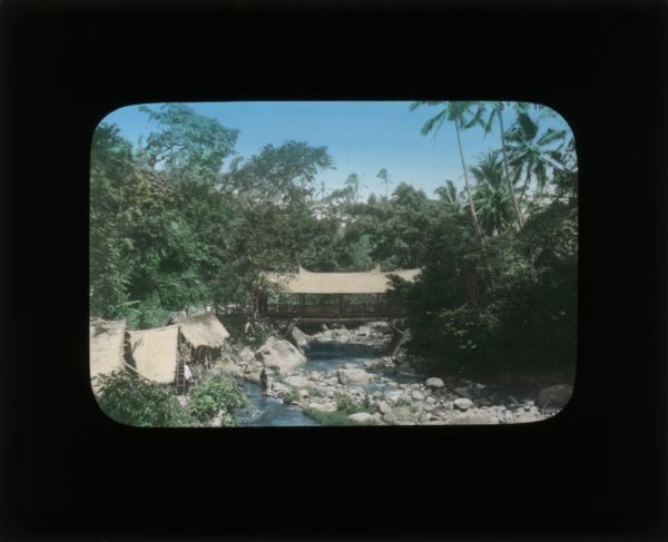 Elevated view of a bridge over a rocky river. In the lower left foreground people are near small, thatched roof buildings, with what appear to be wooden mill wheels beside them, along a smaller stream which forks off before the bridge. In her journal from China, Carrie writes that some of the poorest live in these river-bed dwellings.