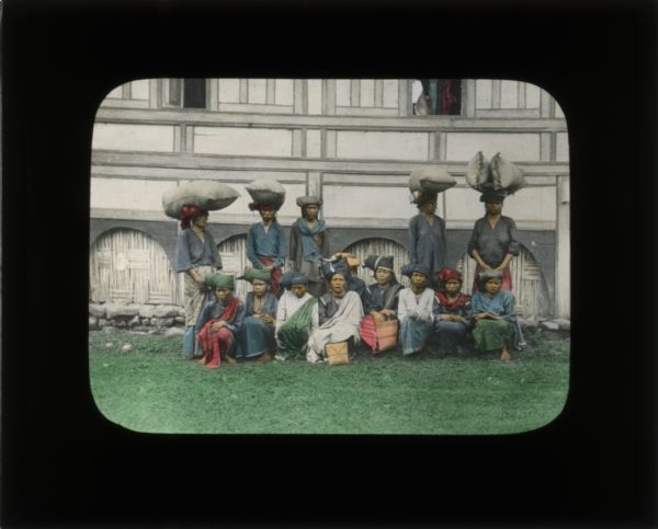 Five women, four of them carrying large sacks of grain on their head, stand behind a group of women sitting on the ground. All of them are wearing head wraps, skirts, and shirts. Behind them is the side of a large building.