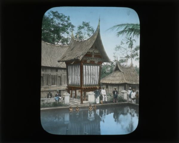 Four children standing waist-deep in a cement pool. Around the pool men and a young child, all wearing head wraps, stand or sit on the cement wall surrounding the pool. Large buildings with thatched roofs are behind them. The smaller building in the middle closest to the pool is supported by stilts, and has decorative weaving or painting under the eaves. There are trees in the far background. The buildings have what Carrie described as the "horn-like" roofs seen in China and the Philippines.