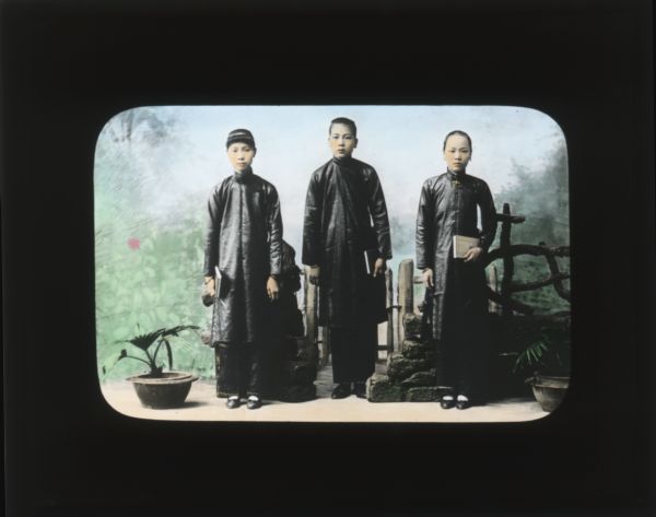 Group portrait in front of a painted backdrop of three boys in black robes each holding a book. The boys stand with two potted plants, and a prop wall of stone and wood. In her journal from Japan and South Korea, Carrie describes the robes worn by students as "black and billowing, over black trousers and with black shoes".