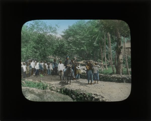 In her journal from Sudan, Carrie describes a tiger hunt which occurred while she was visiting a village. The tiger had been preying on livestock for a couple of days. This slide shows the men returning after the hunt, carrying the tiger on a litter. A group of children is watching in the background on the left.
