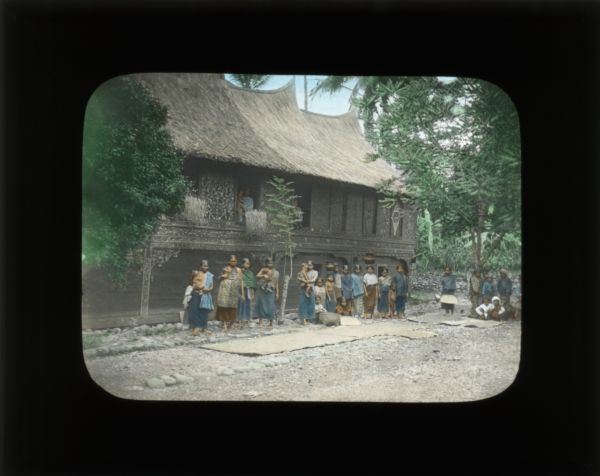 Group of people outside of a village dwelling. The building has the "horn-like" roof which Carrie describes in her journal from China and the Philippines. The people are barefoot. Some are shown with pots on their heads or head wrappings, and most are fully clothed in skirts and blouses.