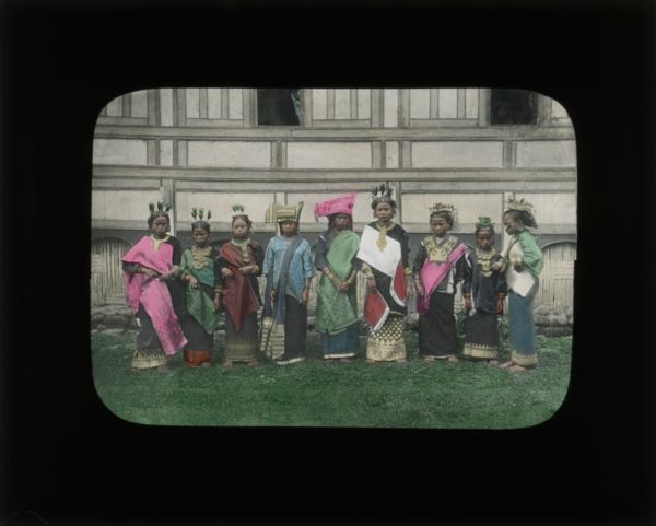 Group portrait of women wearing a variety of head dresses posed in front of the side of a building. They are wearing either black and gold dresses or skirts with blouses, and some are also wearing sashes. Most have gold colored detailing. They are all wearing their hair up and are barefoot.