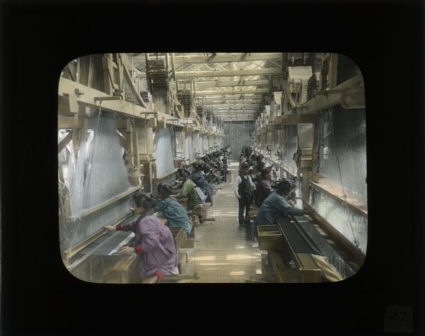 View down aisle of two rows of women workers using looms in a textile factory. A few men stand in the aisle. In her journal from China, Carrie describes the condition of these factories: "The conditions are crowded, the hours are long, the women bent on their work. These, much like plantations, provide wages enough to allow one to live, but not quite so much to as ever live better."