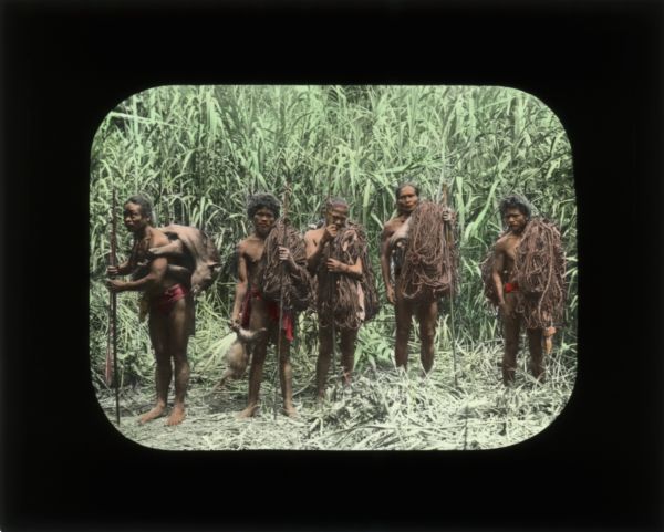 Group portrait of men after a hunt. The man on the left is carrying a Tapir and the man next to him is holding a baby Tapir. Four of the men are holding ropes and some are carrying spears.