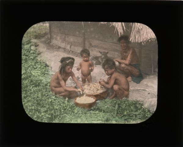 Three children, an adult, and a dog sitting outside of a thatched home. The children appear to be preparing food. All of the children are unclothed. In her journal from Sudan, Carrie writes that "the mothers don't often bother [the children] with clothes."