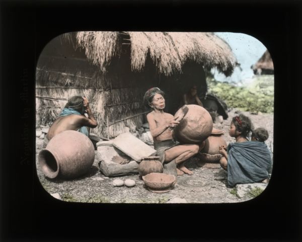 Group of people sitting on the ground outside of a thatched house with pottery.