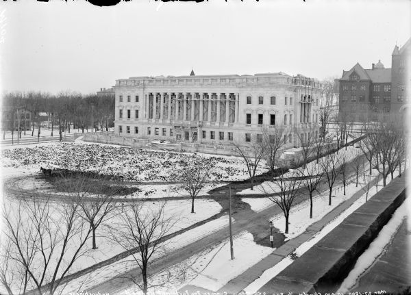 Elevated view over Langdon Street towards the State Historical Society building, which is near completion. Written across the bottom (in reverse): "Mar. 27-1900 10 AM Cloudy 1/2 sec 256 Stop Cramer Iso Inst. not backed Hydrochinone." Men are near a team of horses with a wagon in the snowy yard, and more men are standing on scaffolding near the columns of the building. University of Wisconsin buildings including Science Hall are in the background.