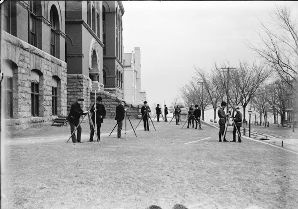 A class of engineering students practicing surveying in front of Science Hall, located on the University of Wisconsin (Madison) campus. Eleven engineering students stand in the grass with their surveying instruments. Lake Mendota is in the background.