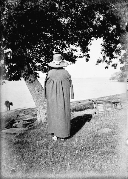 Rear view of an unknown women standing barefoot in the grass near a tree on the shore of a lake. She is wearing a bathing cloak and a straw hat. There is an overturned wheelbarrow on the right, and a cow is wading on the shoreline on the left.