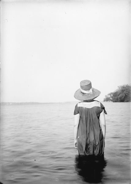 Rear view of an unknown woman standing in a lake wearing a bath dress and a straw hat. Trees are along the shoreline on the right.